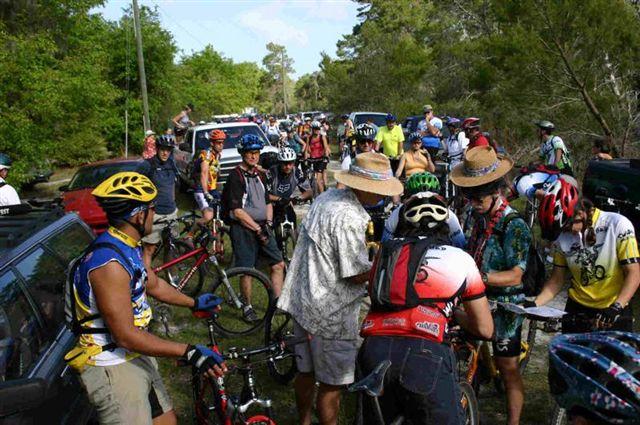 A group of cyclists gathered in a wooded area, preparing for a biking event. Various individuals are wearing helmets and cycling gear, with some bikes propped up nearby. In the background, cars are parked along a dirt path, and trees surround the scene, indicating a natural outdoor setting. The atmosphere appears lively and communal, with participants engaging with one another. Balm Boyette Scrub Preserve mountain bike trail.