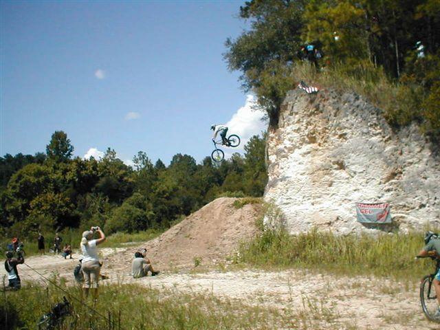 A BMX rider performs a mid-air jump off a dirt ramp, soaring above a low cliff in a wooded area. Spectators are gathered nearby, some taking photos as they watch the action. A banner is visible on the cliffside, and clear blue skies are overhead. Santos mountain bike trail.