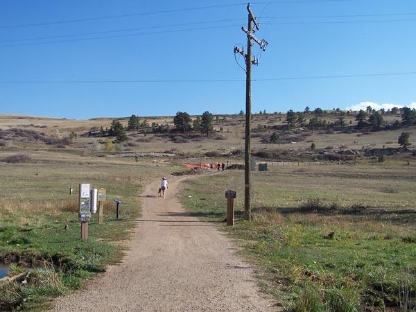 A dirt path leads through a grassy landscape towards a hill with scattered trees under a clear blue sky. A person walks along the path, while signs are visible on either side of the trail. In the background, there are distant figures near a fenced area. A telephone pole stands alongside the path. Marshall Road Open Spaces mountain bike trail.