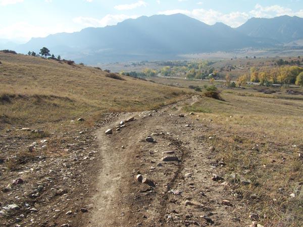 A dirt path winding through grassy terrain, with scattered rocks, leading toward a distant view of mountains under a partly cloudy sky. The scene captures a peaceful, natural landscape with a mix of open fields and hints of autumn foliage in the valley below. Marshall Road Open Spaces mountain bike trail.