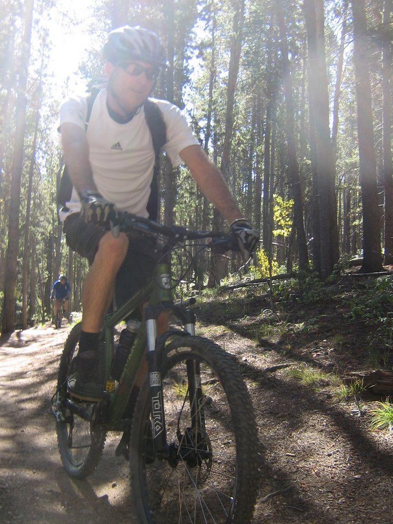A mountain biker riding along a dirt trail through a forest, sunlight filtering through the trees. The cyclist is wearing a helmet, sunglasses, and a white athletic shirt with black shorts. Another biker is visible in the background. Colorado Trail: Kenosha Pass To Breckenridge mountain bike trail.