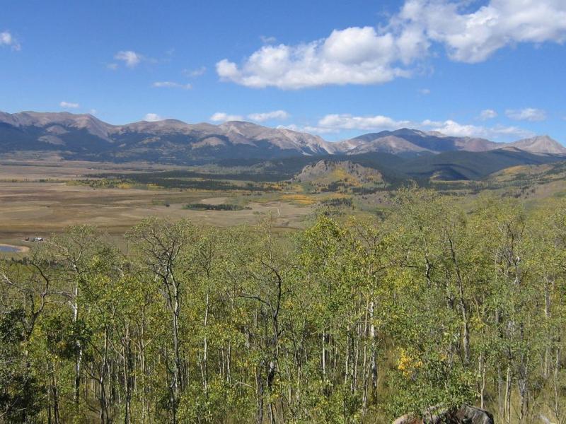 A panoramic view of a mountainous landscape featuring rolling hills and valleys, with a mix of green and yellow foliage in the foreground. The sky is bright blue with scattered white clouds, and the distant mountains are rugged and partially covered in shadows, indicating varying elevations. Colorado Trail: Kenosha Pass To Breckenridge mountain bike trail.