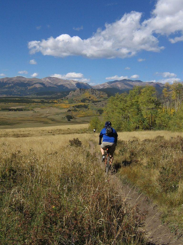 A person biking on a dirt trail through a grassy landscape, with mountains in the background under a blue sky filled with fluffy clouds. Colorado Trail: Kenosha Pass To Breckenridge mountain bike trail.
