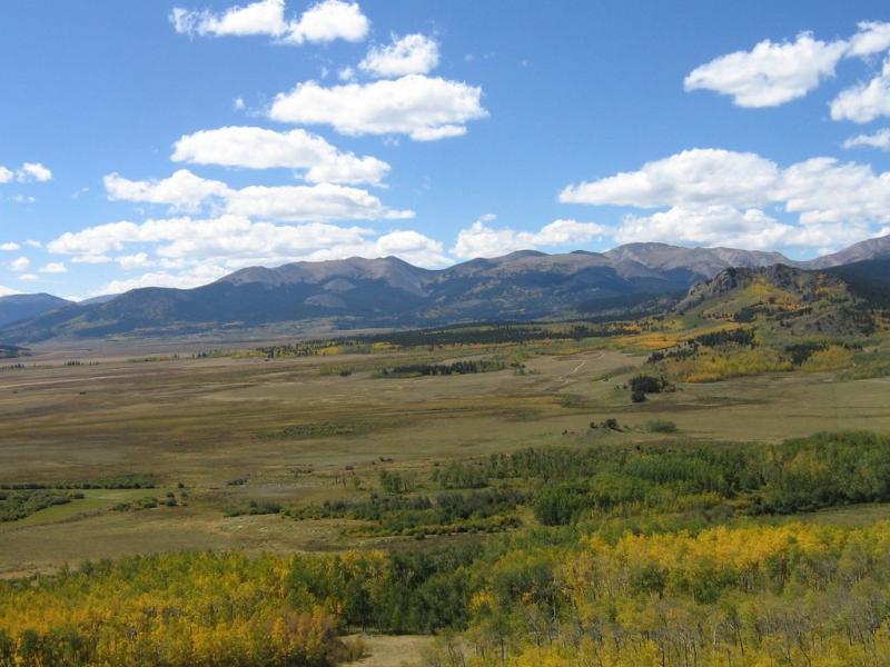 A scenic view of a mountainous landscape with rolling hills and a clear blue sky dotted with clouds. The foreground features vibrant green trees and patches of yellow foliage, indicating the transition of seasons, while the distant mountains are partially covered in lush greenery and rocky outcrops. Colorado Trail: Kenosha Pass To Breckenridge mountain bike trail.