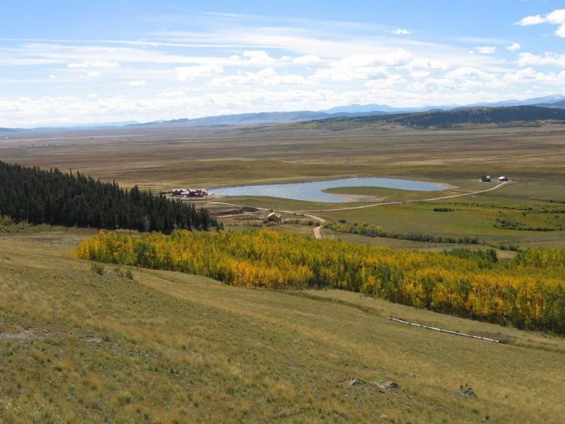 A panoramic view of a serene landscape featuring a small pond surrounded by grassy fields and patches of trees. In the foreground, vibrant yellow and green foliage contrasts against the expansive brown and green terrain. The distant mountains are visible under a blue sky with scattered white clouds, creating a peaceful natural setting. Colorado Trail: Kenosha Pass To Breckenridge mountain bike trail.