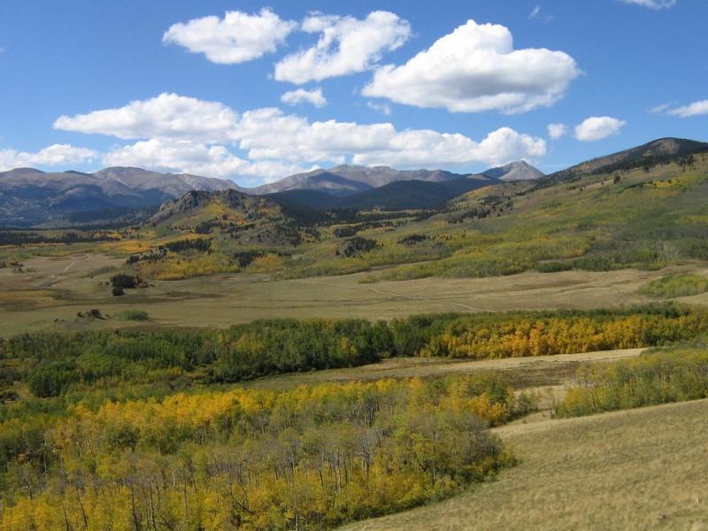 A scenic landscape featuring rolling hills and mountains in the background, adorned with patches of autumn foliage. The foreground showcases a mix of green and yellow trees, while fluffy white clouds drift across a bright blue sky. Colorado Trail: Kenosha Pass To Breckenridge mountain bike trail.
