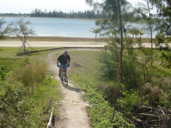 A cyclist riding a mountain bike along a dirt path surrounded by greenery, with a lake visible in the background and a clear sky overhead. Amelia Earhart Park mountain bike trail.