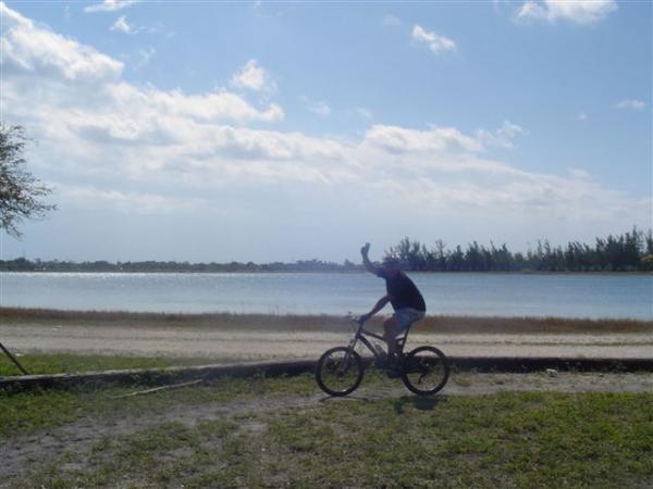 A person riding a bicycle along a path near a calm body of water, waving their hand. The scene is set against a backdrop of blue skies with scattered clouds and greenery along the shoreline. Amelia Earhart Park mountain bike trail.