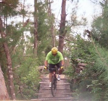 A cyclist wearing a bright green shirt and a yellow helmet rides a mountain bike down a wooden trail surrounded by trees and greenery. Amelia Earhart Park mountain bike trail.