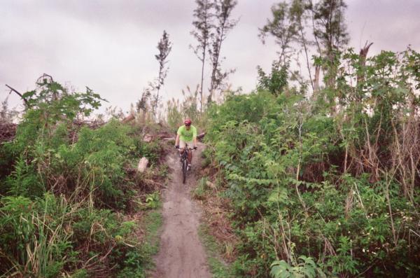 A cyclist riding down a narrow, grassy trail surrounded by dense vegetation and trees, under a cloudy sky. The rider is wearing a bright green shirt and a helmet. Amelia Earhart Park mountain bike trail.