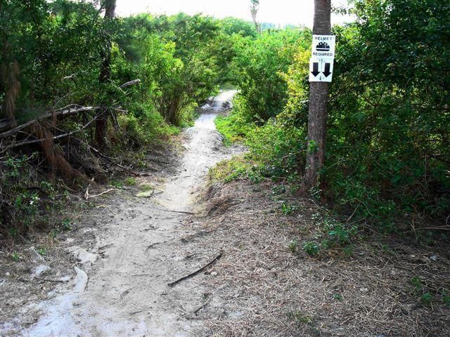 A narrow dirt path surrounded by dense greenery, leading into a wooded area. On a nearby tree, there is a sign indicating that a helmet is required. The trail appears to be used for biking or hiking. Amelia Earhart Park mountain bike trail.
