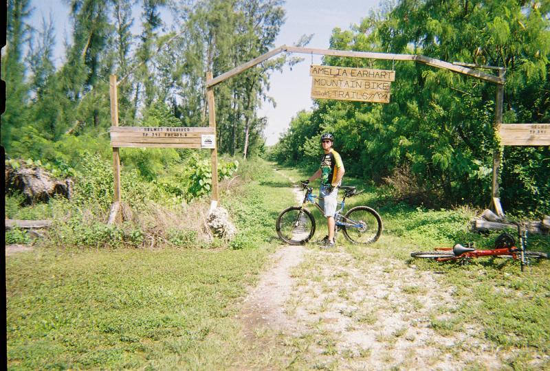 A cyclist stands next to a mountain bike in front of the Amelia Earhart Mountain Bike Trails entrance, with a wooden archway overhead. The trail is surrounded by lush greenery and trees, indicating a natural outdoor setting. A red bike is partially visible on the ground nearby. Amelia Earhart Park mountain bike trail.