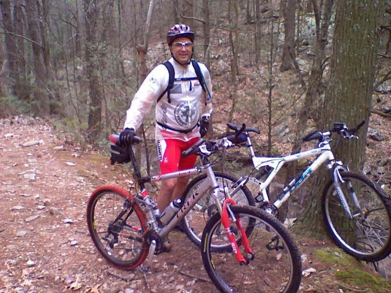 A man in cycling gear stands next to two mountain bikes on a wooded trail. He is wearing a helmet and a light jacket with a logo, and has a backpack on. The background features trees and rocky terrain, indicating an outdoor cycling adventure. Elizabeth Furnace mountain bike trail.