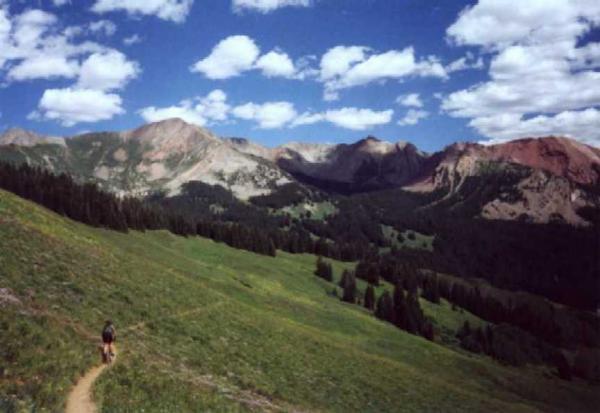 A hiker walking along a dirt trail through a green meadow, surrounded by mountains under a blue sky with fluffy white clouds. Trail 401 mountain bike trail.