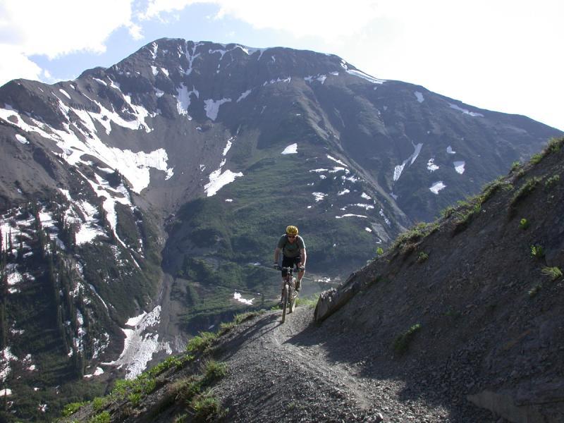 A mountain biker navigates a narrow gravel path on a hillside, surrounded by steep mountains and patches of snow. The sky is clear with a few clouds, highlighting the rugged terrain and lush greenery below. Trail 401 mountain bike trail.