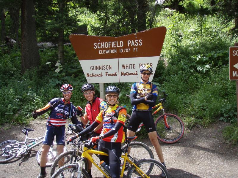 Four cyclists pose in front of the Schofield Pass sign, which indicates an elevation of 10,707 feet. The background features lush greenery typical of a forested area, and two bicycles are parked nearby. The cyclists are dressed in colorful cycling gear, conveying a sense of camaraderie and accomplishment. Trail 401 mountain bike trail.