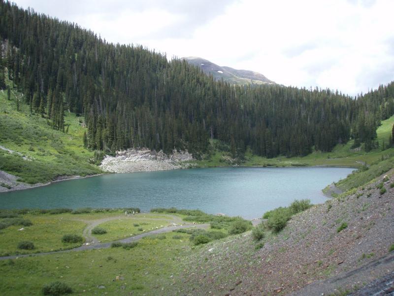 A serene mountain lake surrounded by lush green forests and gentle slopes. The still water reflects the cloudy sky, while a winding path leads along the shoreline. Rocky terrain and patches of wildflowers create a picturesque natural landscape. Trail 401 mountain bike trail.