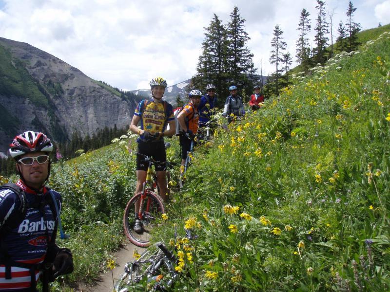 A group of mountain bikers pause along a trail surrounded by vibrant wildflowers and greenery, with mountain scenery in the background. The cyclists, wearing helmets and cycling gear, are gathered on a dirt path, showcasing a mix of excited expressions and relaxed poses. Trail 401 mountain bike trail.