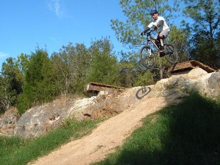 A mountain biker performing a jump off a dirt ramp, surrounded by trees and rocky terrain under a clear blue sky. Santos mountain bike trail.
