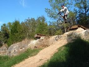 A cyclist performing a jump on a mountain bike over a dirt ramp, surrounded by trees and blue skies. Santos mountain bike trail.