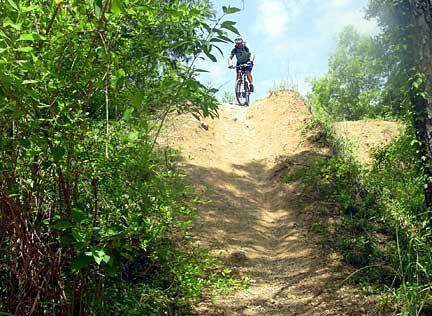 A mountain biker performing a jump off a dirt ramp in a wooded area, surrounded by greenery and under a partly cloudy sky. Santos mountain bike trail.