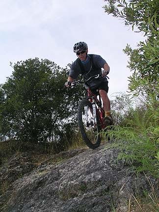 A person riding a mountain bike down a rocky slope surrounded by greenery. The cyclist is wearing a helmet and sunglasses, demonstrating an adventurous outdoor activity. Santos mountain bike trail.
