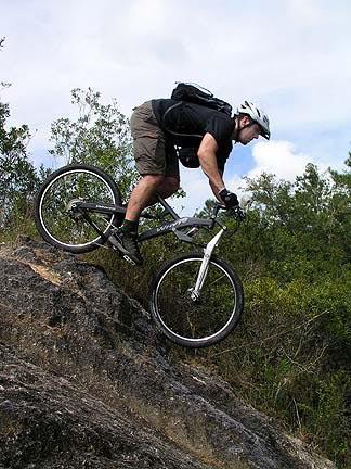 A mountain biker descending a rocky slope, wearing a helmet and backpack, surrounded by greenery and blue skies. Santos mountain bike trail.