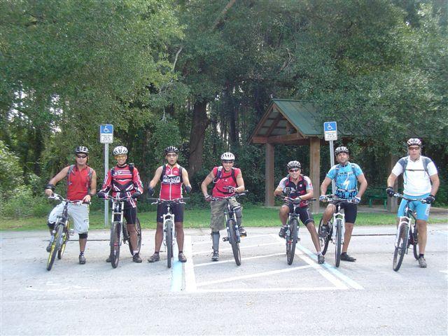 A group of seven mountain bikers, wearing helmets and biking attire, pose with their bicycles in a park setting. They are lined up on a pathway, with trees and a shelter visible in the background. The cyclists exhibit a range of styles and equipment, showcasing a community of biking enthusiasts ready for an outdoor adventure. Santos mountain bike trail.