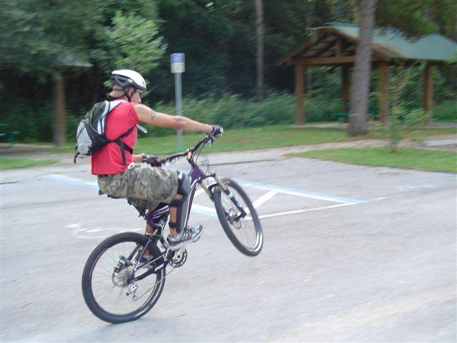 A person wearing a helmet and a red shirt performs a wheelie on a mountain bike in a park setting. The background features trees and a gazebo, with a parking area visible in the foreground. Santos mountain bike trail.