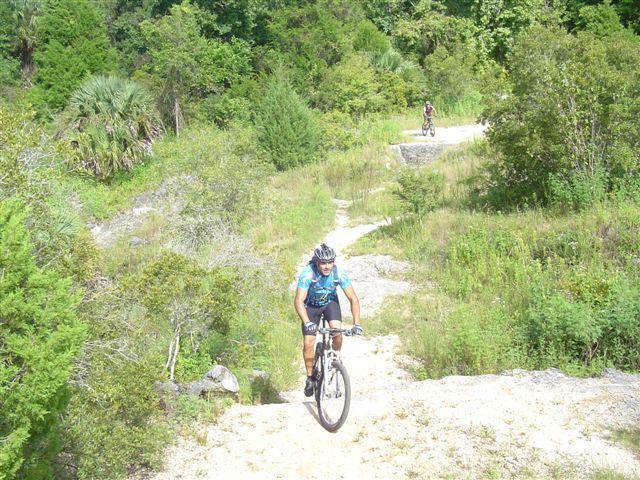 Two mountain bikers riding on a dirt trail surrounded by lush greenery. The foreground features a cyclist in a blue and black outfit navigating a slope, while another cyclist can be seen in the background on a higher elevation of the trail. The landscape includes dense shrubs and trees, indicating a natural, outdoor setting. Santos mountain bike trail.