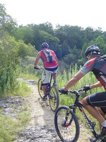 Two mountain bikers riding on a rocky trail surrounded by greenery. One biker is dressed in a red shirt and white shorts, while the other is in a black and red jersey. The trail is narrow and leads through a lush landscape. Santos mountain bike trail.