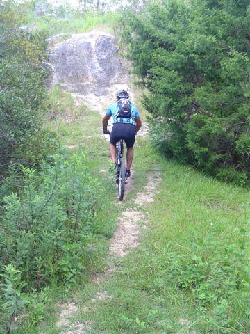 A mountain biker wearing a blue jersey and helmet rides along a narrow dirt trail surrounded by greenery and rocks. Santos mountain bike trail.
