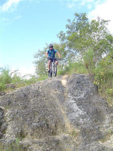 A mountain biker descends a rocky outcrop surrounded by greenery under a blue sky. Santos mountain bike trail.