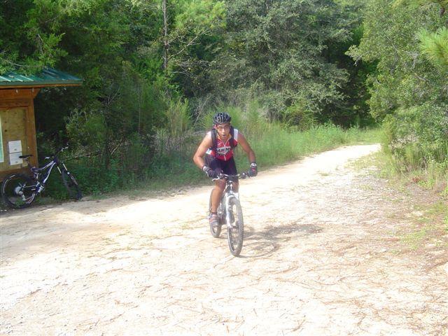 A person riding a mountain bike on a dirt path surrounded by greenery. In the background, another bike is resting against a shelter, and the area is illuminated by natural light. Santos mountain bike trail.