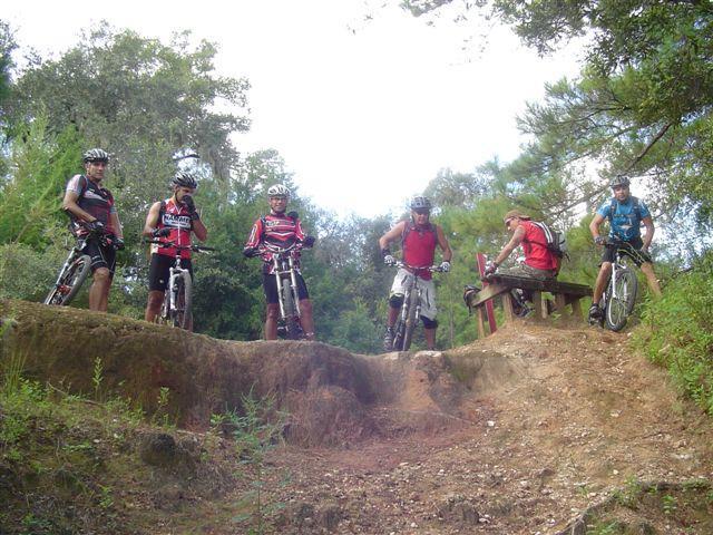 A group of five mountain bikers in biking gear stands on an elevated dirt path surrounded by trees. One biker is seated on a bench, while the others are positioned near their bikes, looking down the slope. The scene captures an outdoor biking adventure, showcasing the natural terrain and sporty attire. Santos mountain bike trail.