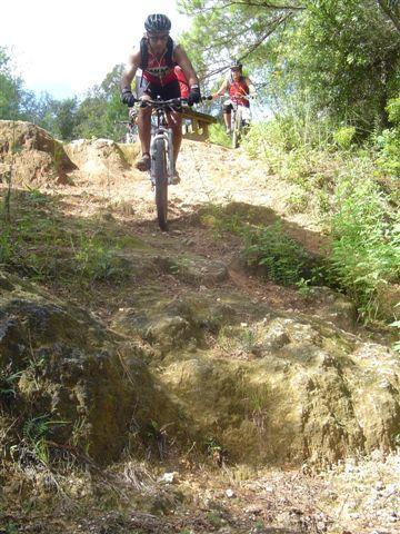 A mountain biker navigating a rocky trail in a lush, green environment, with another cyclist visible in the background. The scene captures the thrill of outdoor biking in nature. Santos mountain bike trail.