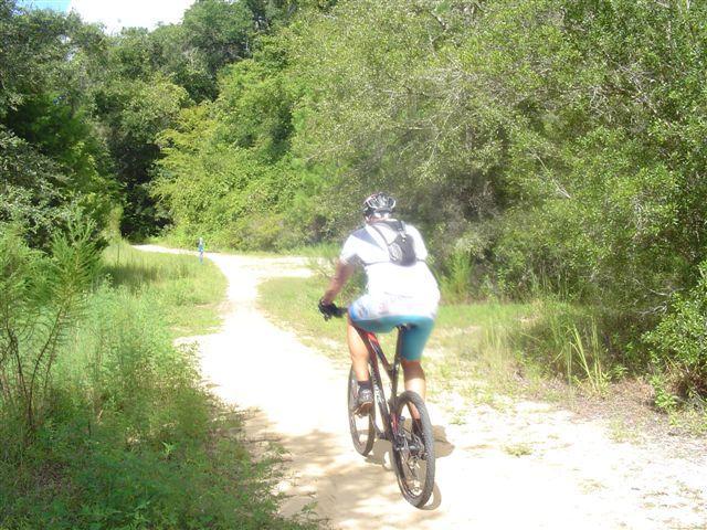 A person riding a mountain bike on a dirt trail surrounded by lush greenery on both sides, with another cyclist visible in the distance. The scene depicts a sunny day in a natural outdoor setting. Santos mountain bike trail.