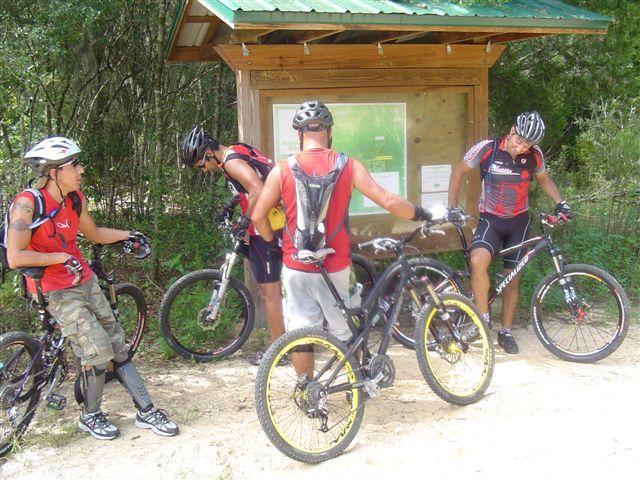 A group of four mountain bikers, wearing helmets and cycling gear, gather around a trail map at a wooded location. The cyclists are examining the map for directions while their bikes are propped nearby. The setting is sunny, with lush vegetation in the background. Santos mountain bike trail.