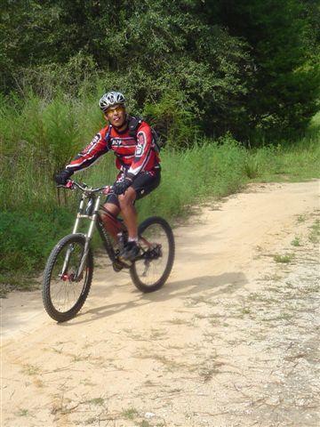 A person wearing a colorful cycling jersey and helmet rides a mountain bike along a sandy trail surrounded by greenery. Santos mountain bike trail.