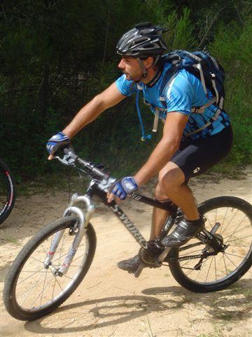 A cyclist in a blue jersey and helmet rides a mountain bike along a dirt trail, surrounded by greenery. The rider is focused and leaning forward as they navigate the path. Santos mountain bike trail.