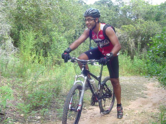 A person riding a mountain bike on a dirt trail surrounded by greenery, wearing a helmet and athletic gear, and smiling as they navigate the path. Santos mountain bike trail.