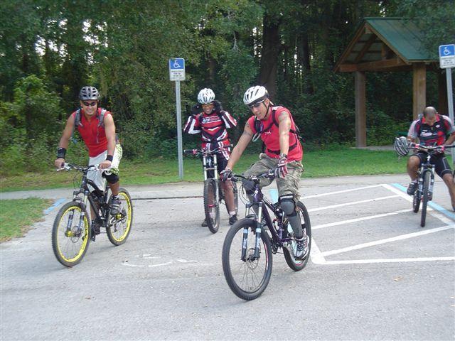 Four men in cycling gear are gathered at a bike parking area surrounded by trees. They are on mountain bikes, with two wearing red jerseys and one in a black and red outfit. The scene captures a moment before they begin riding, showcasing their helmets and protective gear. A sign indicating accessible parking is visible in the background. Santos mountain bike trail.