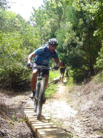 A mountain biker riding on a wooden bridge along a narrow trail surrounded by lush greenery, with another rider visible in the background. Santos mountain bike trail.
