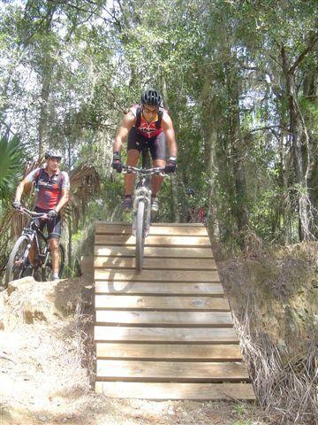 A mountain biker descends a wooden ramp, navigating a trail surrounded by lush greenery. A second cyclist watches from the side, while additional riders can be seen in the background, enjoying the outdoor biking experience. Santos mountain bike trail.