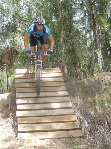 A mountain biker riding a wooden ramp in a forested area, with trees and foliage surrounding the scene. The cyclist is wearing a helmet and athletic attire, focused on navigating the ramp. Santos mountain bike trail.