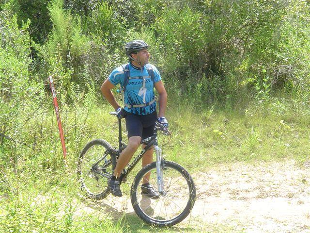 A male mountain biker wearing a blue and black jersey and a helmet is standing next to his bicycle on a path surrounded by green vegetation. He appears to be looking off into the distance, contemplating his next move on the trail. Santos mountain bike trail.