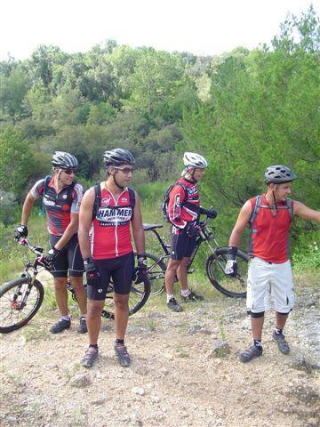 Four mountain bikers in sportswear stand on a dirt path surrounded by greenery. They wear helmets and are holding their bikes while looking in different directions. The scene captures a moment of camaraderie and exploration in an outdoor setting. Santos mountain bike trail.