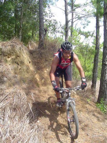 A person riding a mountain bike on a dirt trail surrounded by trees. The rider is wearing a black helmet and a red and black cycling outfit, navigating a sloped section of the path. Sunlight filters through the trees, creating a natural outdoor setting. Santos mountain bike trail.