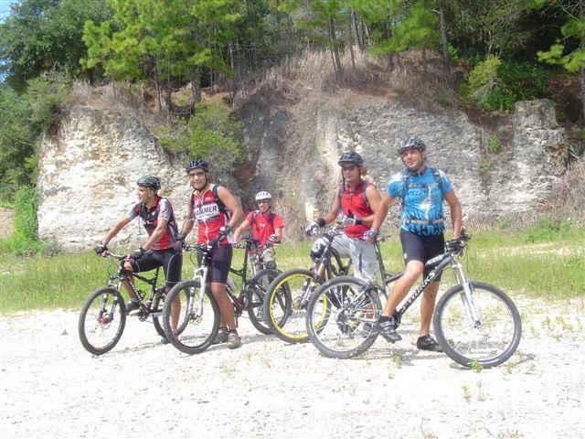 Five mountain bikers posing with their bikes on a dirt path surrounded by greenery and rocky terrain. They are wearing helmets and athletic clothing, with a backdrop of trees and steep rock formations. Santos mountain bike trail.