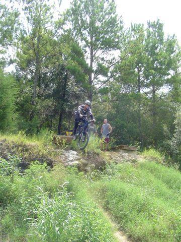 A mountain biker leaps off a small ramp on a trail surrounded by greenery, while a person stands nearby watching. Sunlight filters through the trees, creating a vibrant outdoor scene. Santos mountain bike trail.